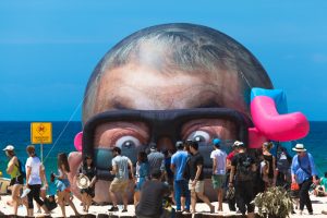 Crowds walking past a large inflatable head sculpture on Bondi Beach during Sculpture by the Sea.